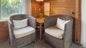Two wicker chairs with white cushions, a small table, a lamp, and a framed picture on a wooden wall.
