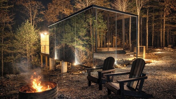 Une cabane entièrement vitrée, avec un foyer et deux chaises à l’extérieur.