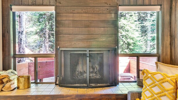 A corner fireplace with a screen, wooden walls, and large windows with a view of trees.
