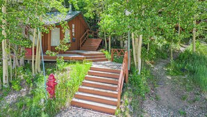 A wooden cabin with a staircase leading to a porch, surrounded by trees and greenery.