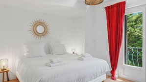 A bedroom with a bed, a bedside table, a lamp, a window with a red curtain, and a view of greenery outside. - Bardelours 1 – Charming house with swimming pool in the heart of the Pic Sa (Saint-Bauzille-de-Montmel)