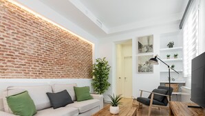 Un salon moderne avec un mur d’accent en briques, un canapé blanc agrémenté de coussins verts et noirs, une table basse en bois, un fauteuil noir et un téléviseur posé sur un meuble en bois.