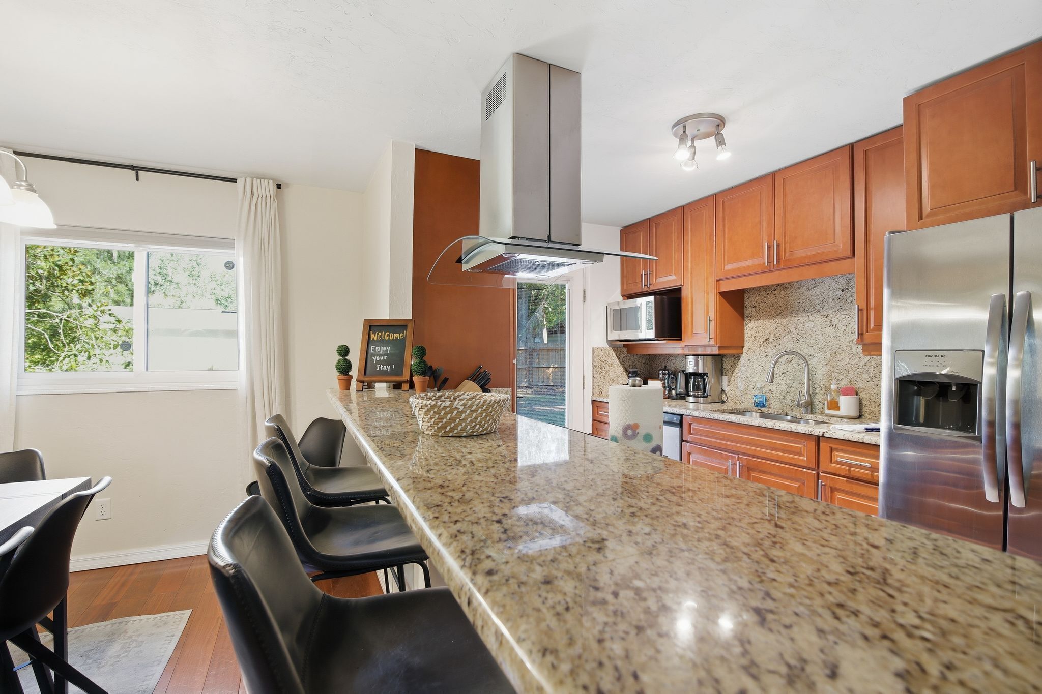A modern kitchen with granite countertops, stainless steel appliances, wooden cabinets, and a bar area with bar stools.