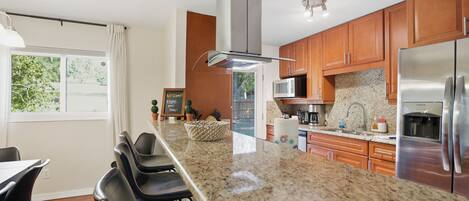 A modern kitchen with granite countertops, stainless steel appliances, wooden cabinets, and a bar area with bar stools.