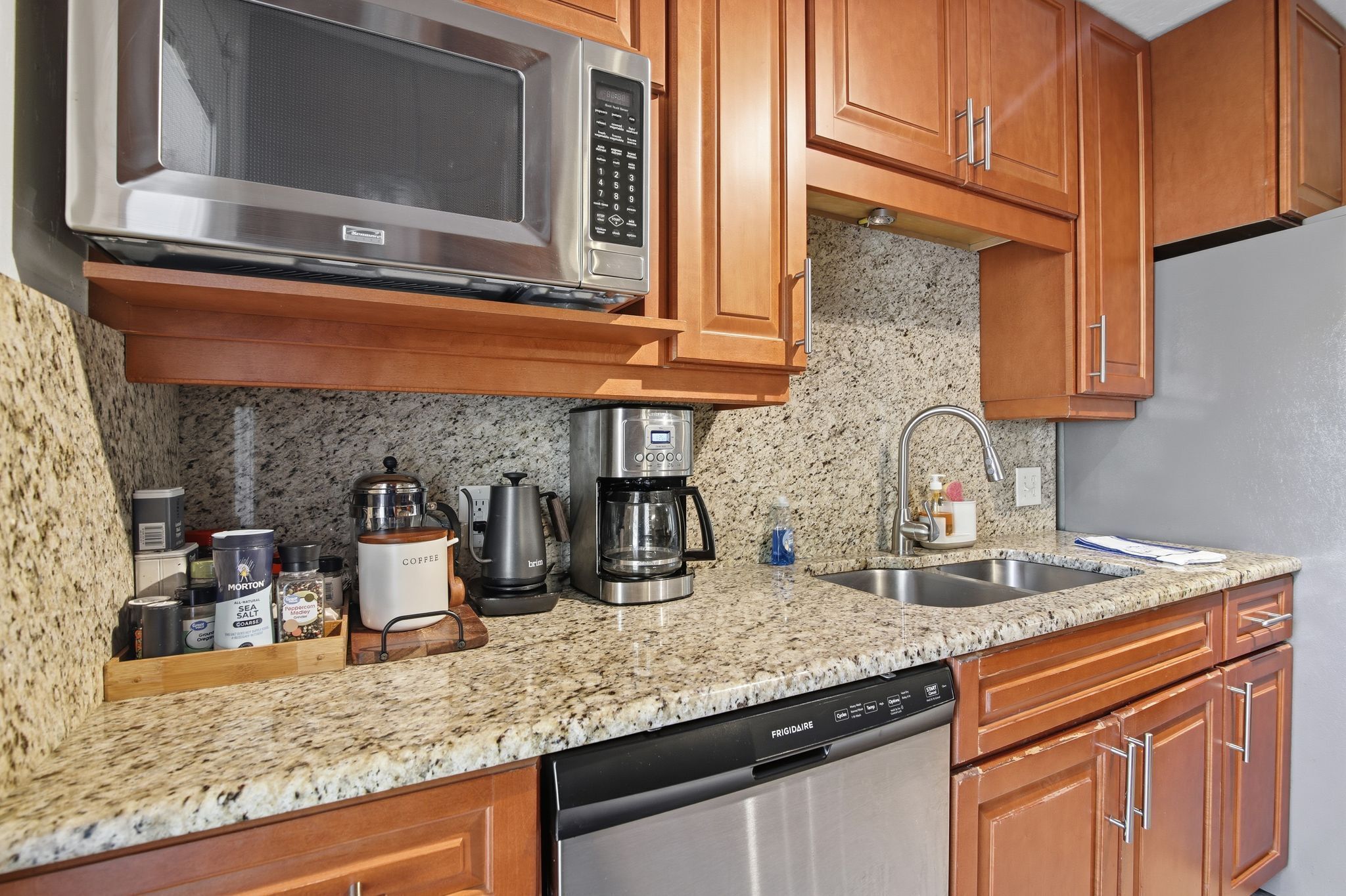 A kitchen with wooden cabinets, a granite countertop, a stainless steel microwave, a coffee maker, and a dishwasher.