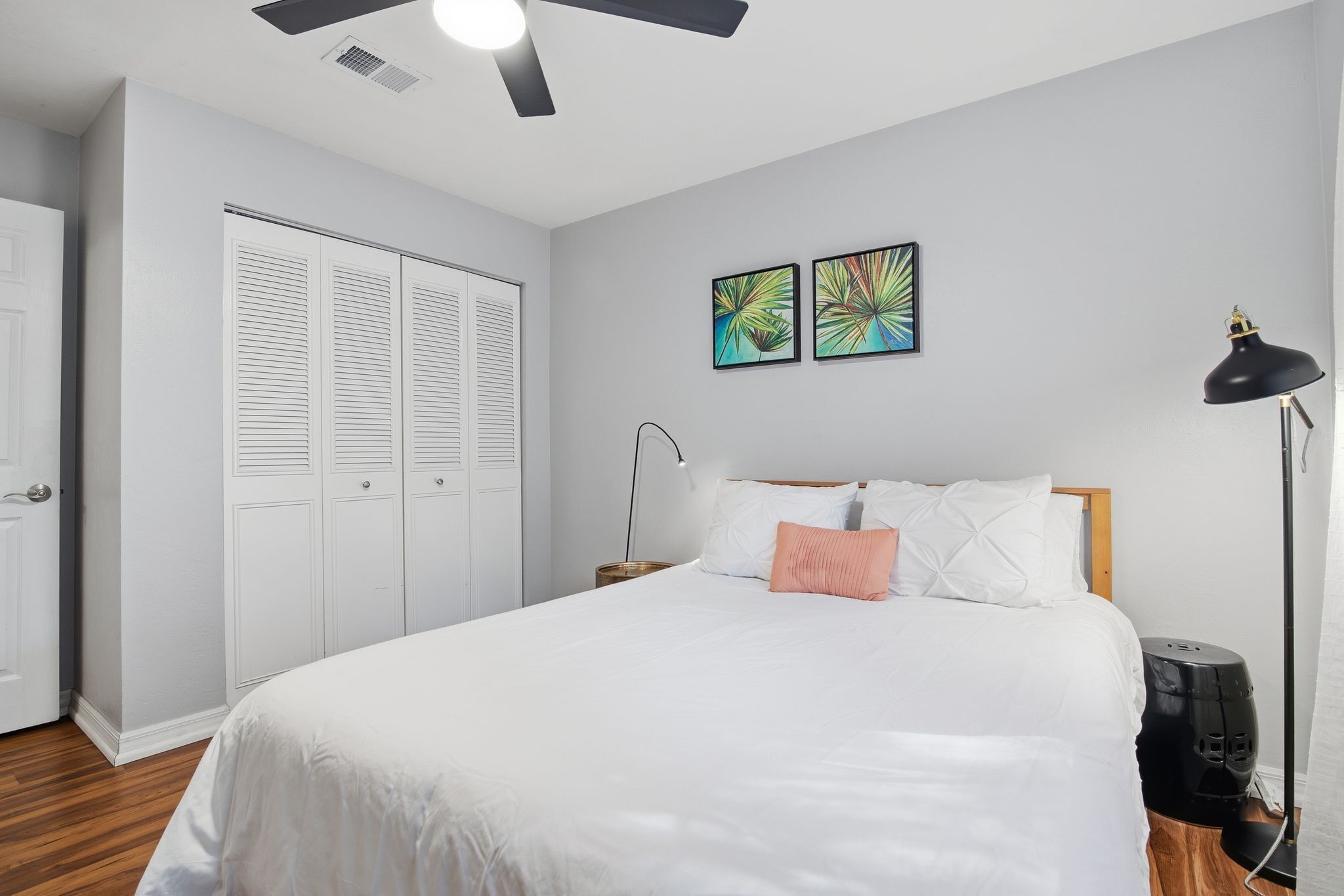 A bedroom with a bed, white walls, a ceiling fan, two wall art pieces, a nightstand, a lamp, and a black speaker.