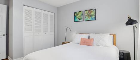 A bedroom with a bed, white walls, a ceiling fan, two wall art pieces, a nightstand, a lamp, and a black speaker.