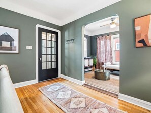 A hallway with a black door, a patterned rug, and a view into a living room with a couch and curtains. - Hot Tub - Game Room - Professional Design-Backyard (University Heights)