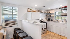 Una cocina moderna con armarios blancos, una isla central, taburetes de bar, un ventanal con persianas y una cafetera.