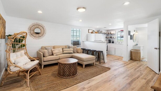 Un salón amplio con un sofá grande en forma de L, una mesa de centro redonda y una silla de mimbre. En el fondo, se ve una cocina con armarios blancos y una zona de comedor con taburetes.
