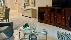 Un salon moderne avec une table basse en verre, des canapés en cuir, un téléviseur, des meubles en bois et des œuvres d’art encadrées accrochées au mur.