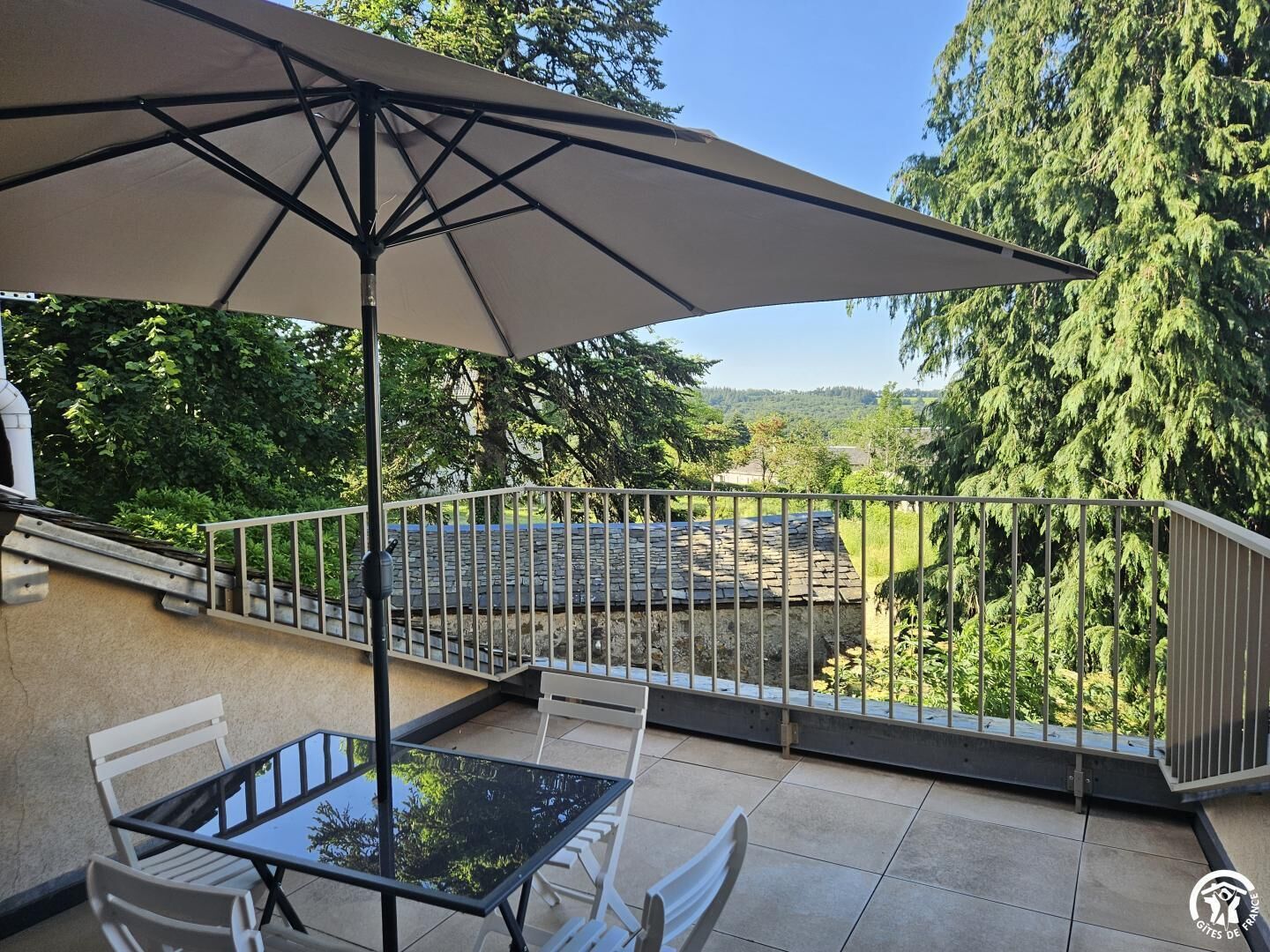 A patio with a table and chairs, a large umbrella, and a view of a stone roof and trees.