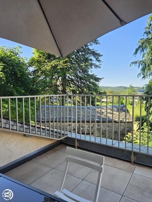 Un balcon doté d’une rambarde en métal, d’un sol carrelé et offrant une vue sur un bâtiment en pierre et sur la verdure.