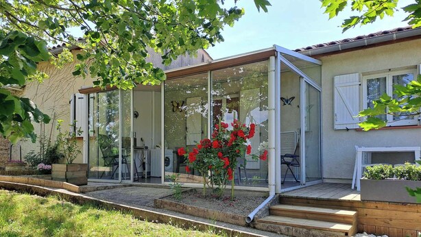 Une maison moderne dotée d’un porche vitré, d’une terrasse en bois et d’un jardin agrémenté de fleurs rouges.