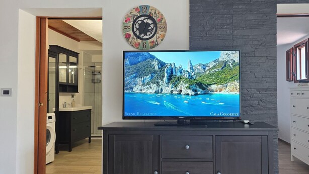 A flat-screen TV on a dark wooden cabinet with a scenic coastal image on the screen. - Porto Frailis Apartment (Arbatax)