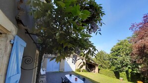 A covered outdoor patio with a table and chairs, surrounded by greenery and a building with a blue door. - Vors - GAILLAC - Gîte (GAILLAC)