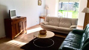 A living room with a sofa, a television on a wooden cabinet, a lamp, and a view of a green landscape through a window.