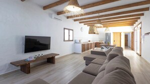 A modern living room with a large flat-screen TV, a wooden bench, and a sofa. There is a kitchen area in the background with wooden cabinets and a dining table.
