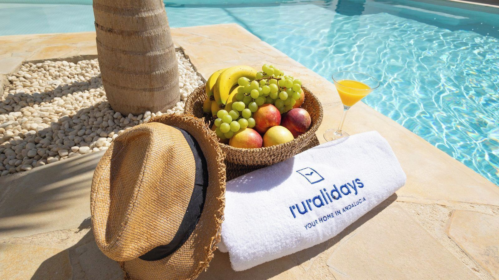 Escena junto a la piscina con una cesta de mimbre llena de fruta, un sombrero de paja y una toalla con un logotipo.