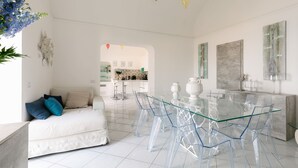 A modern living room with a glass dining table, white sofa, and clear chairs. There is a kitchen area in the background with bar stools. - Villa Tirrena by My Villa Collection (Positano)