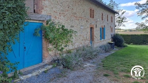 Un bâtiment rustique en pierre, avec une porte et des volets bleus, entouré de verdure et bordé d’un chemin de gravier.