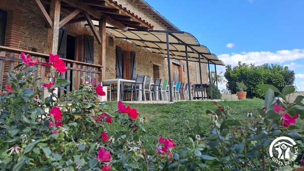 Un bâtiment rustique en pierre, doté d’un patio couvert, comprenant un espace salon extérieur et un jardin agrémenté de fleurs roses.