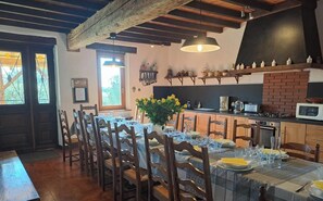 A dining area with a long table set for a meal, wooden chairs, a vase of yellow flowers, and a kitchen with a brick oven in the background.