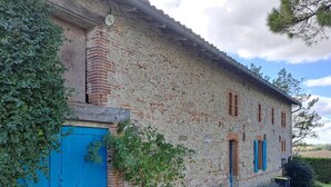 Un edificio rústico de piedra con una puerta y contraventanas azules, rodeado de follaje y un sendero de grava.