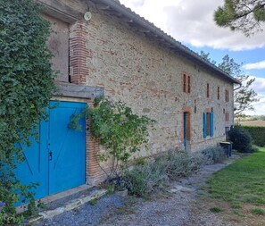 Un edificio rústico de piedra con una puerta y contraventanas azules, rodeado de follaje y un sendero de grava.