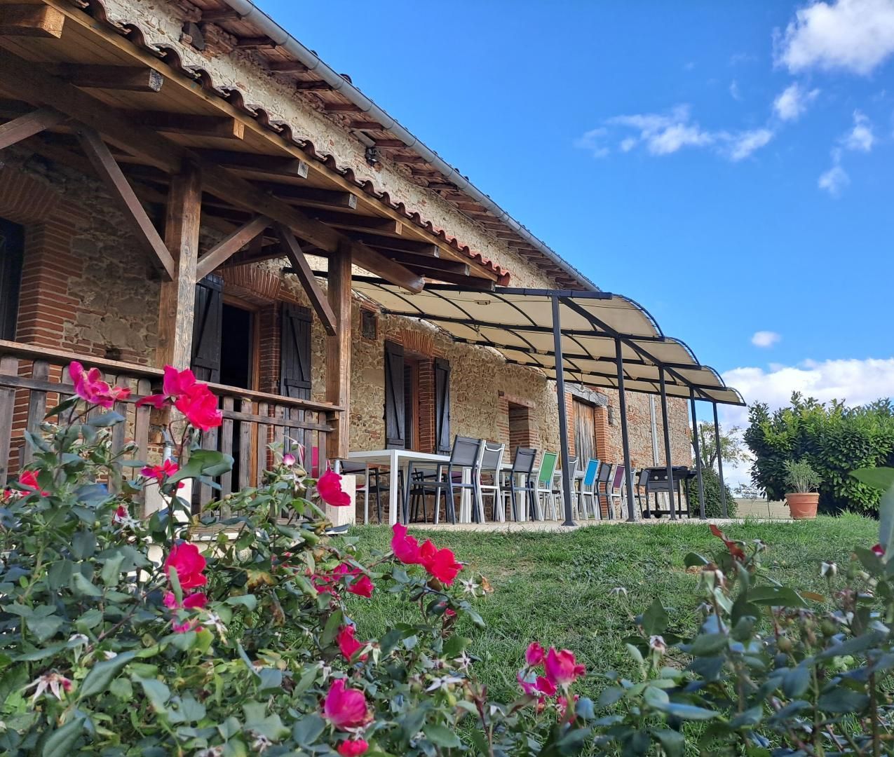 A rustic stone building with a covered patio area, featuring outdoor seating and a garden with pink flowers.