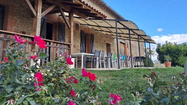 Un edificio rústico de piedra con un porche cubierto, que incluye asientos exteriores y un jardín con flores rosadas.