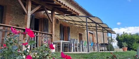 A rustic stone building with a covered patio area, featuring outdoor seating and a garden with pink flowers.