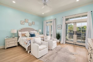 A bedroom with a bed, two white armchairs, a nightstand, a dresser, a large window with curtains, and a ceiling fan.