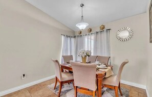 Un comedor con una mesa de madera, cuatro sillas, un jarrón con flores, una araña y un espejo decorativo en la pared.