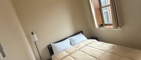 Un dormitorio con una cama, una ventana con cortinas, una lámpara de noche y un enchufe.