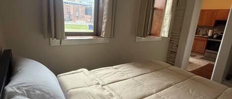 Un dormitorio con una cama grande, dos ventanas y vistas a un edificio de ladrillo.