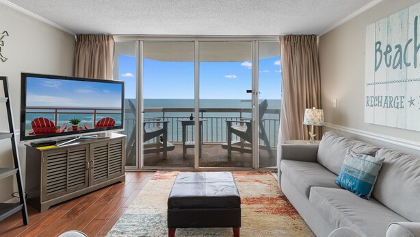 A living room with a television, a sofa, a coffee table, and a view of the ocean through a sliding glass door.