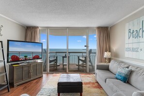 A living room with a television, a sofa, a coffee table, and a view of the ocean through a sliding glass door.