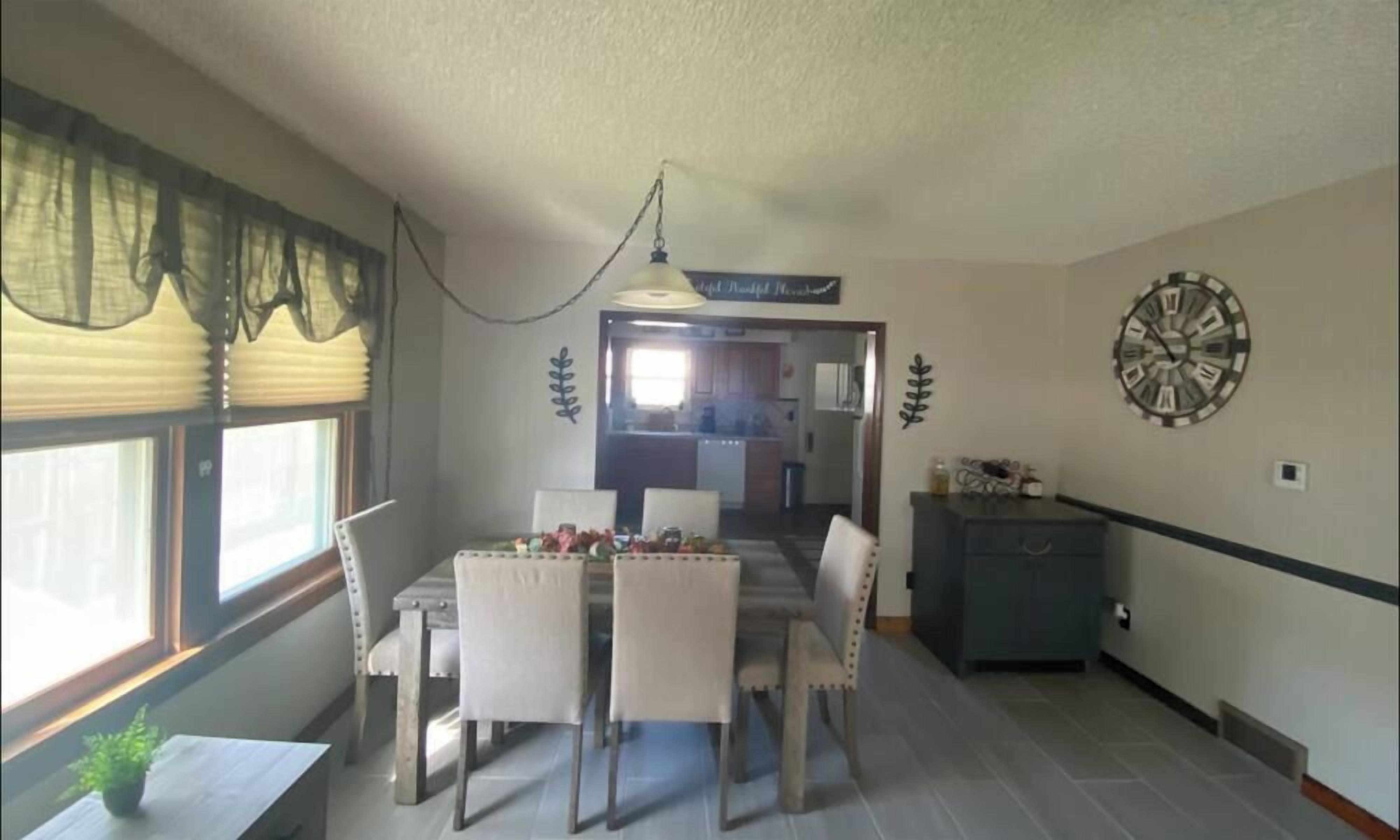 Un comedor con una mesa y sillas, un reloj en la pared y una ventana con persianas.