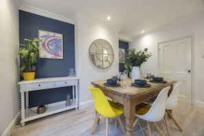 Un coin repas avec une table en bois dressée pour quatre personnes, des chaises blanches, une chaise jaune et une console blanche agrémentée d’une plante.