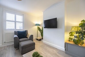 A living room with a grey sofa, a small ottoman, a floor lamp, a large plant, and a flat-screen TV mounted on the wall. - Spacious 3-Bed Period Home Near Bath Road (Cheltenham)