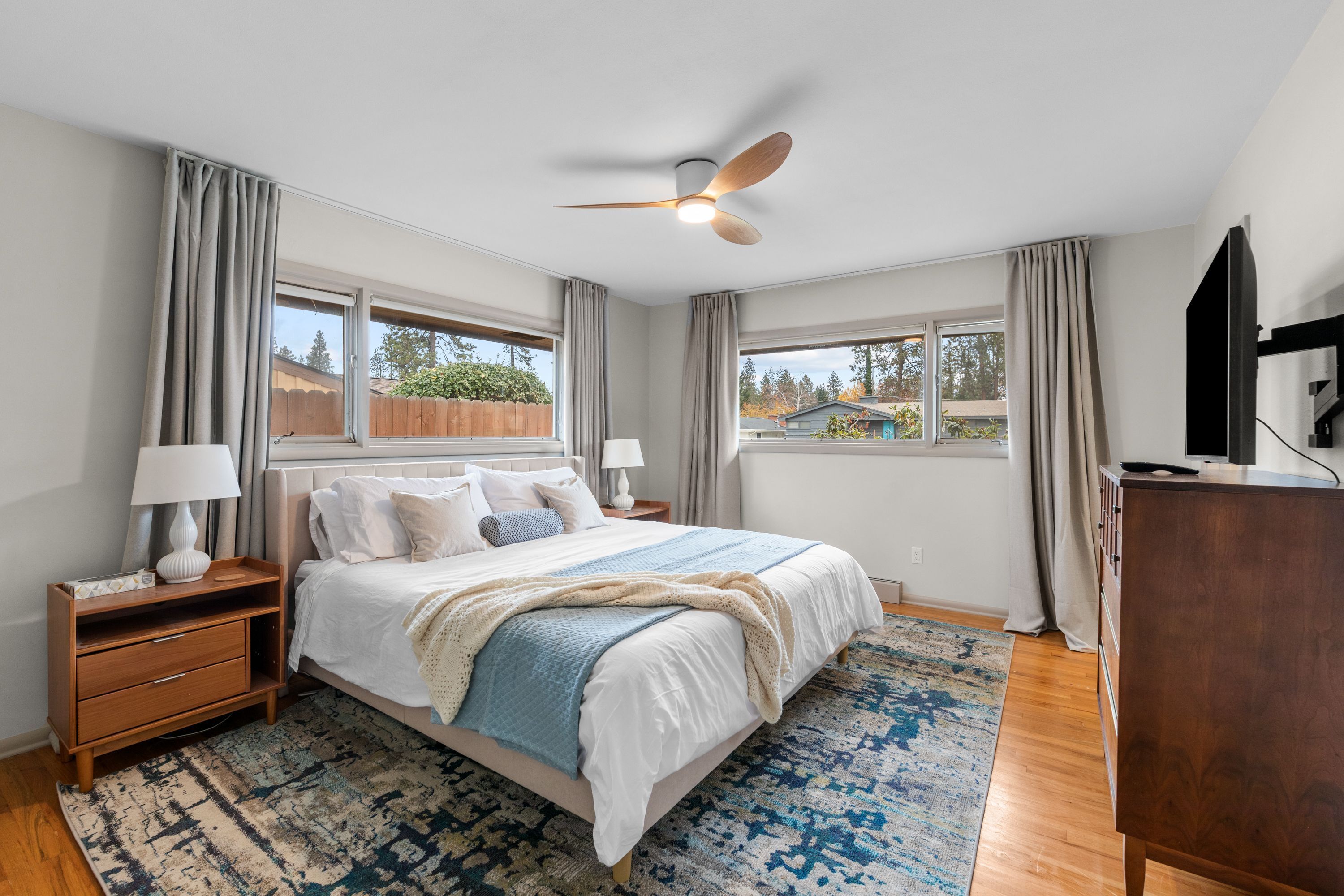 Une chambre à coucher comprenant un lit, des tables de chevet, une commode, un ventilateur de plafond et de grandes fenêtres.