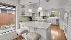 A modern kitchen with white cabinets, a large island with bar stools, pendant lighting, and a view of the backyard. - Stylish South Hill Getaway | Bar & Game Room (Spokane)