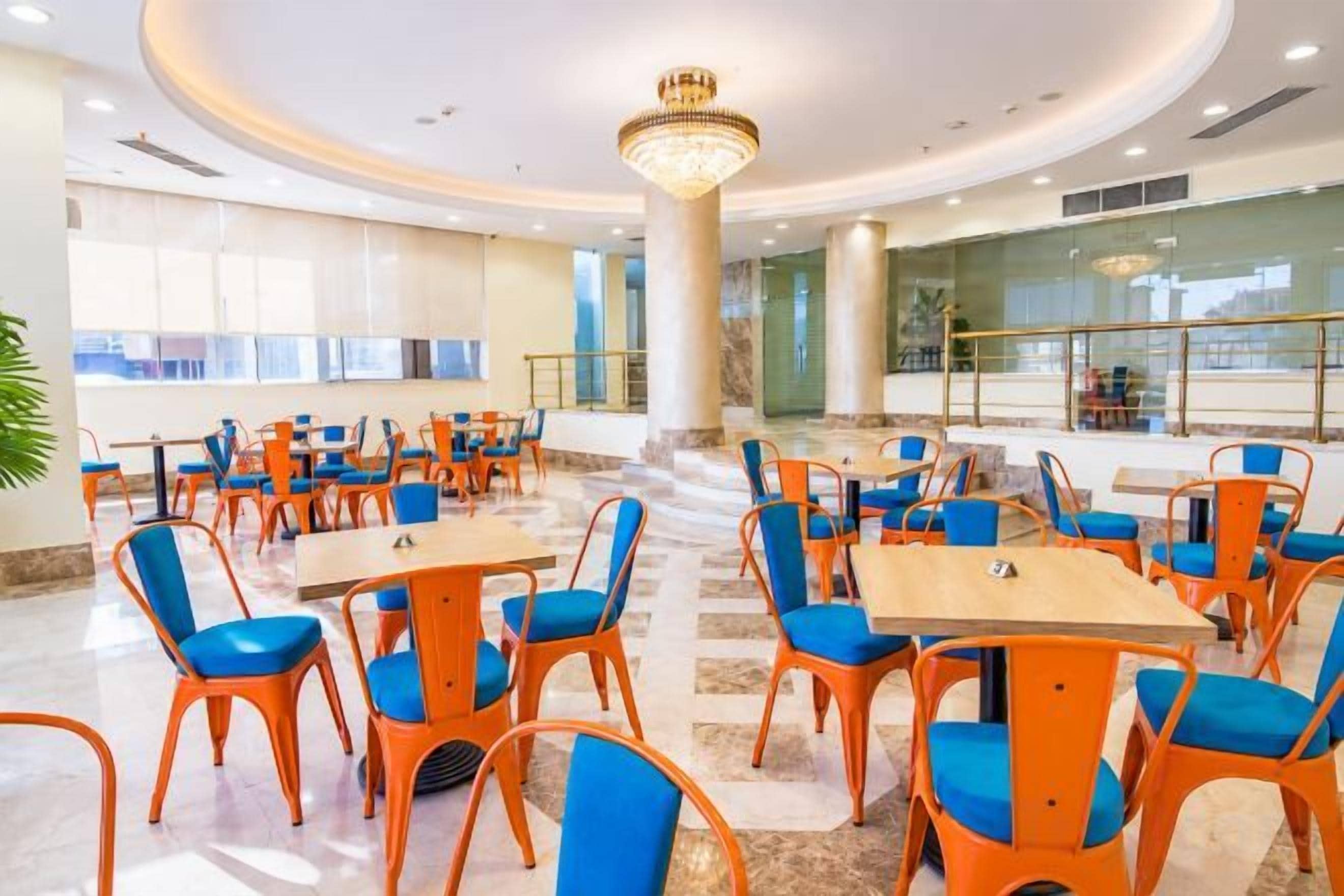 Comedor de hotel con mesas redondas y sillas color naranja, una lámpara de araña y vistas de la ciudad a través de amplias ventanas.