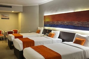 Habitación de hotel con tres camas, cada una con ropa blanca y detalles naranja, un cuadro abstracto grande en la pared y un escritorio pequeño con una silla.
