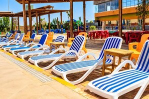 A row of sun loungers with attached tables, arranged in a covered outdoor area with a modern building in the background. - Gloria Aqua Park (Aswan)