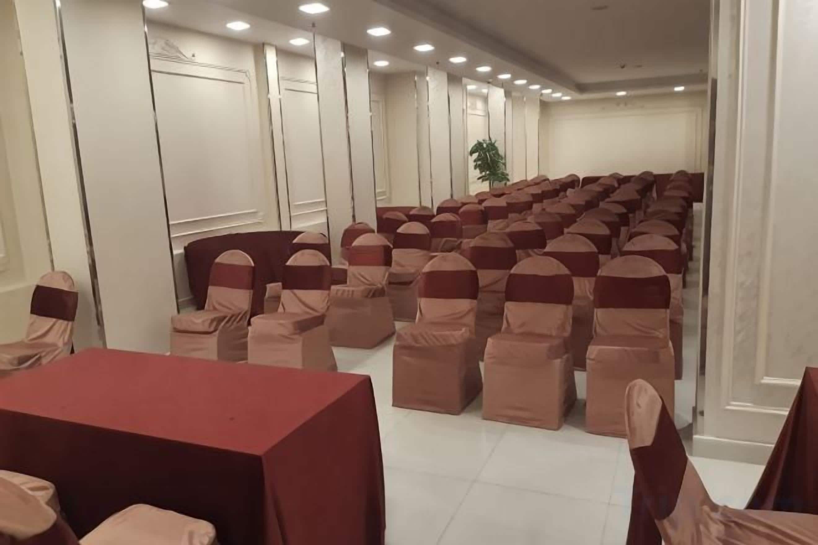 Une salle de conférence avec des rangées de chaises et de tables, des murs blancs et un plafond doté d’un éclairage encastré.