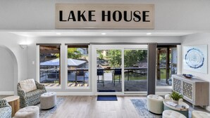 A modern interior with a sign reading "LAKE HOUSE," featuring a seating area with ottomans, a coffee table, and a view of an outdoor patio with a table and chairs.