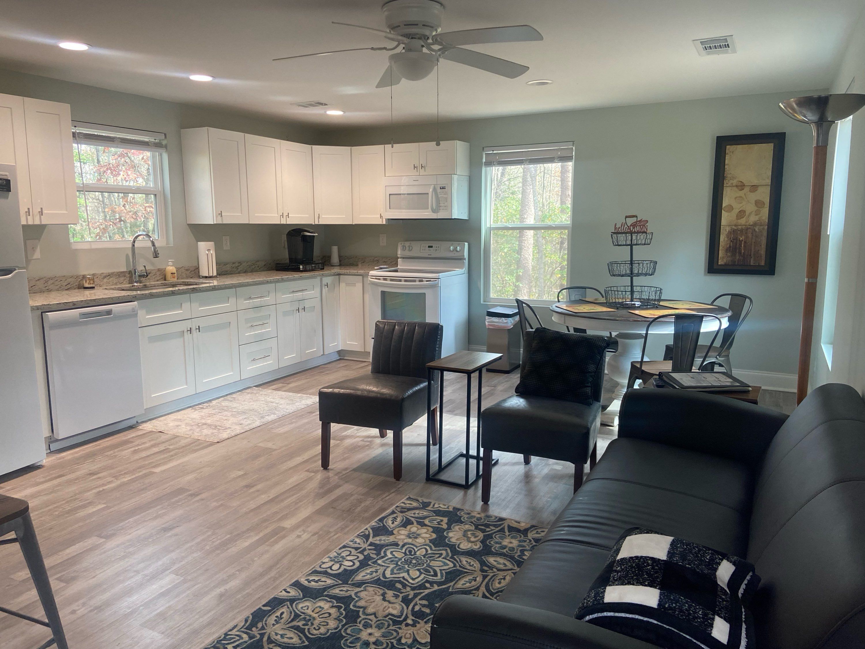 A modern kitchen with white cabinets, a dining area with a table and chairs, and a living room with a black leather sofa and a patterned rug.