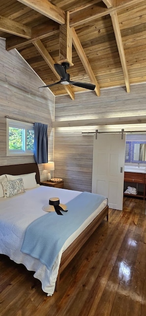 Une chambre avec un plafond en bois, un lit recouvert d’une couverture bleue, une table de chevet avec une lampe et une fenêtre avec des rideaux.